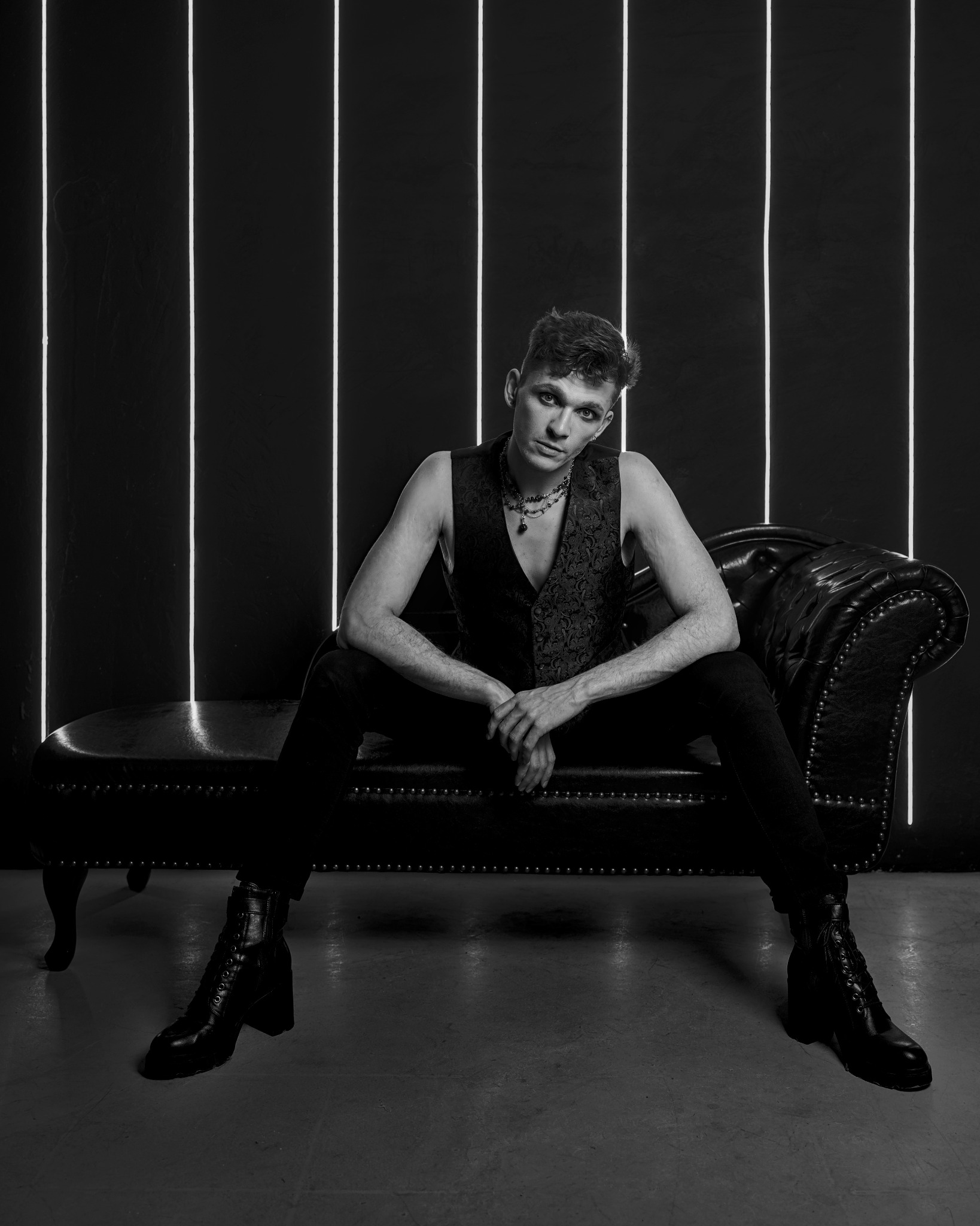 Czarno-biały portret młodego mężczyzny w kamizelce, siedzącego na skórzanej kanapie na tle ściany z neonowymi lampami. Ujęcie centralne, ciemna tonacja.