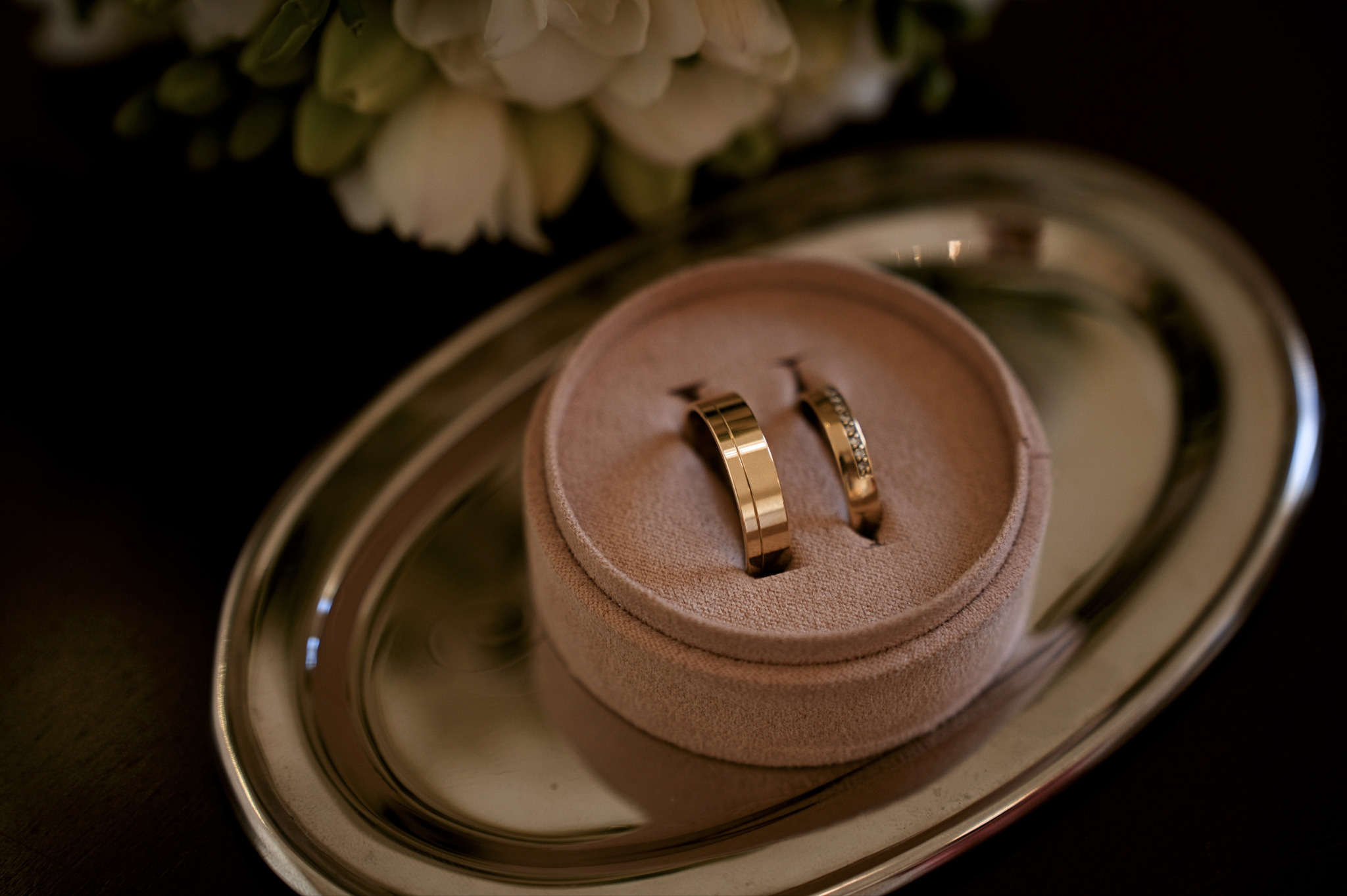 Dwa złote pierścionki ślubne, jeden z brylantami, leżące w beżowym pudełku na srebrnej tacy. W tle rozmyte białe kwiaty. Delikatne światło.