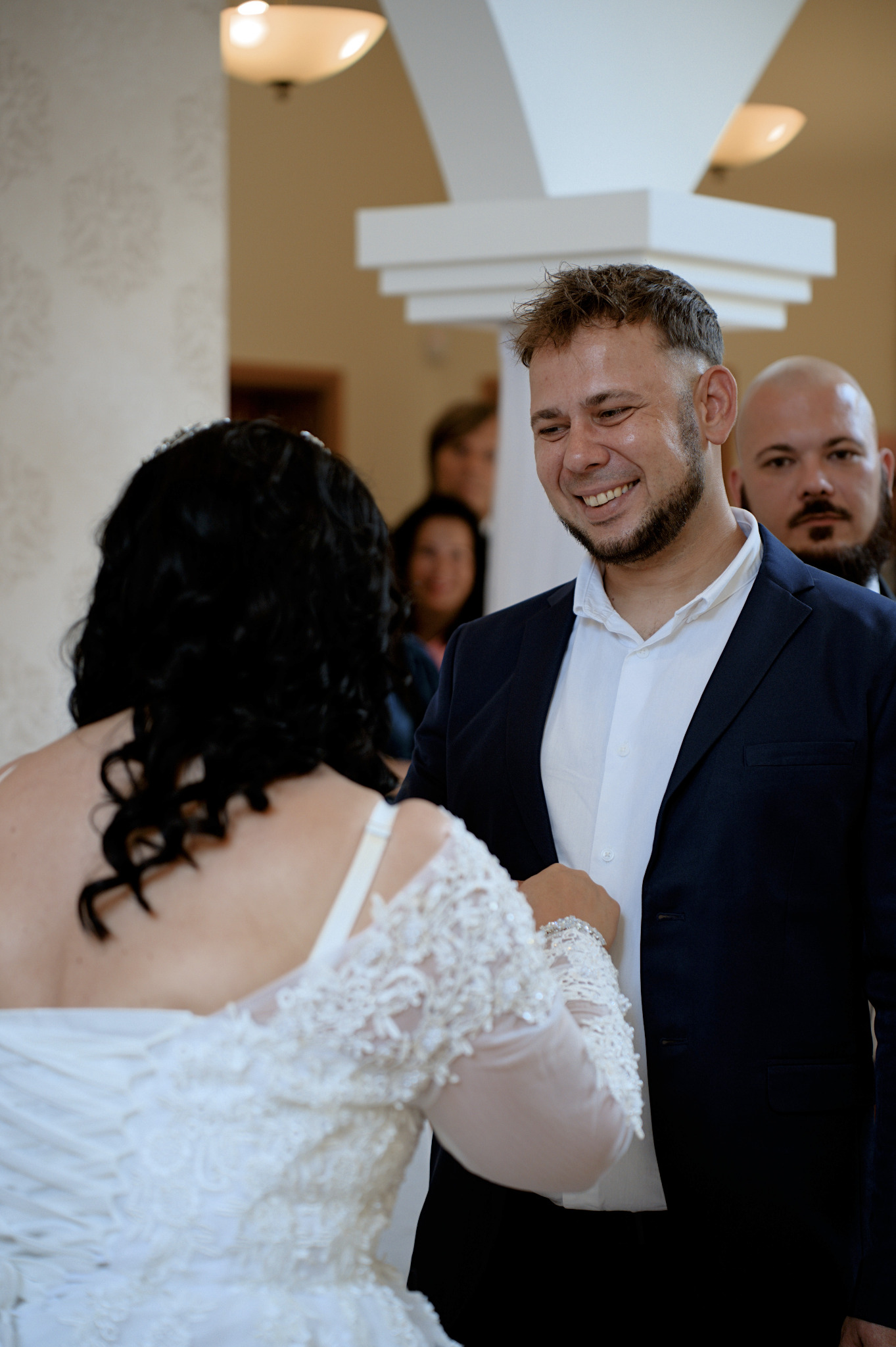 Uśmiechnięty pan młody w granatowym garniturze spogląda na pannę młodą w białej sukni z koronkowymi zdobieniami. W tle goście weselni i kolumna.