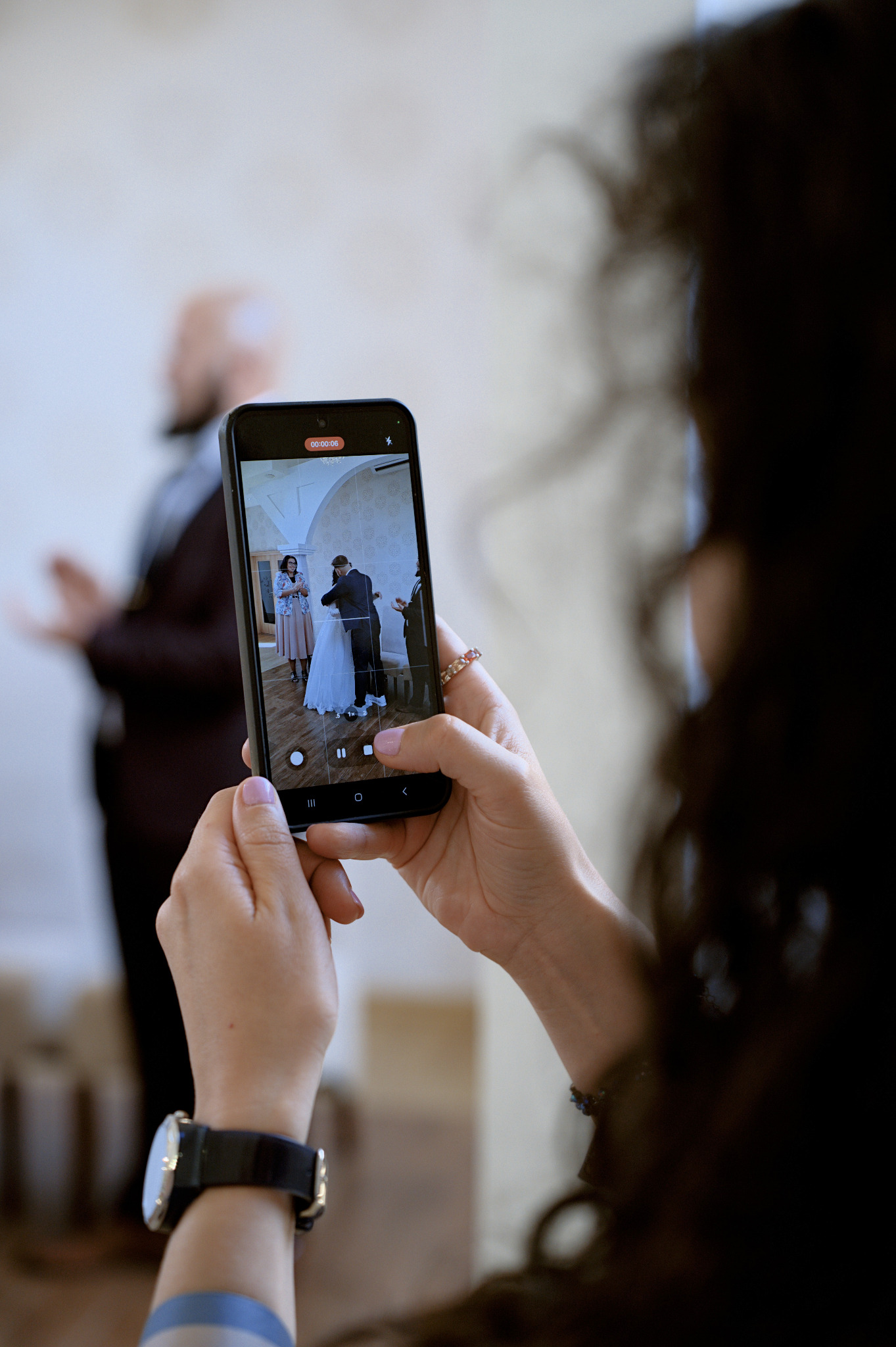 Osoba filmuje telefonem parę młodą podczas ceremonii ślubnej. W tle widoczny mężczyzna. Na ekranie telefonu widoczny kadr z uroczystości.