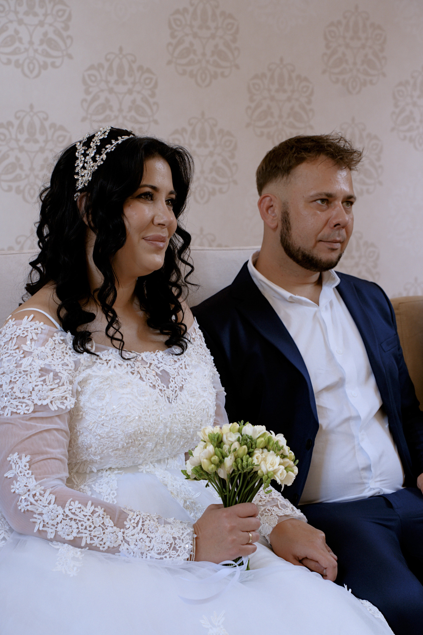 Para młoda siedzi obok siebie: panna młoda w koronkowej sukni trzyma bukiet frezji, obok pan młody w granatowym garniturze i białej koszuli. W tle wzorzysta tapeta.