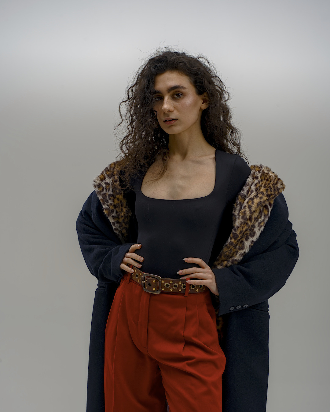 Portret kobiety z kręconymi włosami w czarnym body, czerwonych spodniach i płaszczu z lamparcim futrem, pozującej na jasnym tle. Ujęcie studyjne.