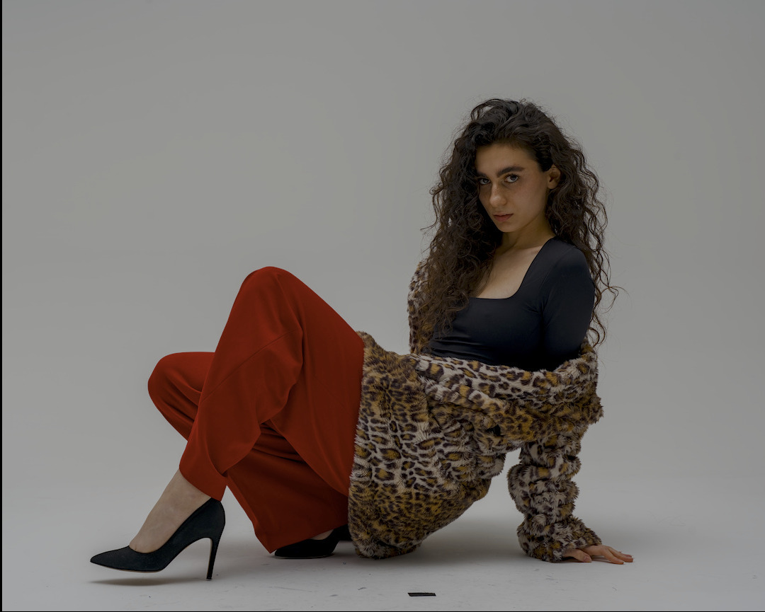 Kobieta z kręconymi włosami w czarnej bluzce, czerwonych spodniach i futrze w panterkę, siedząca na podłodze w szpilkach, pozująca do zdjęcia w studio.