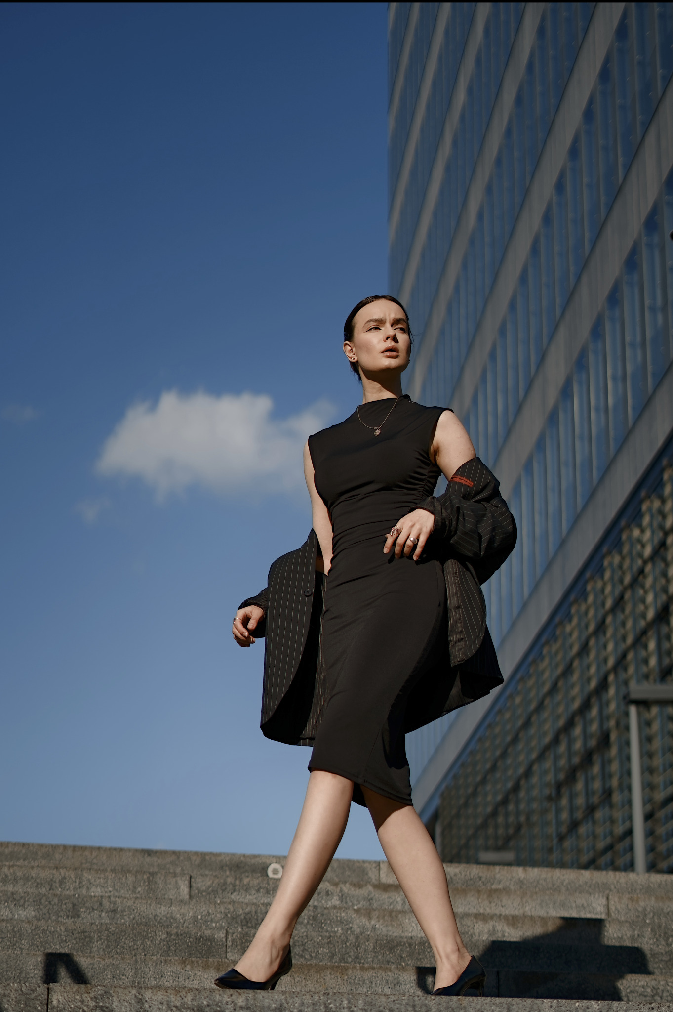 Kobieta w czarnej sukience i marynarce na schodach, z nowoczesnym budynkiem w tle. Ujęcie z dołu, podkreślające sylwetkę i architekturę. Słoneczny dzień.