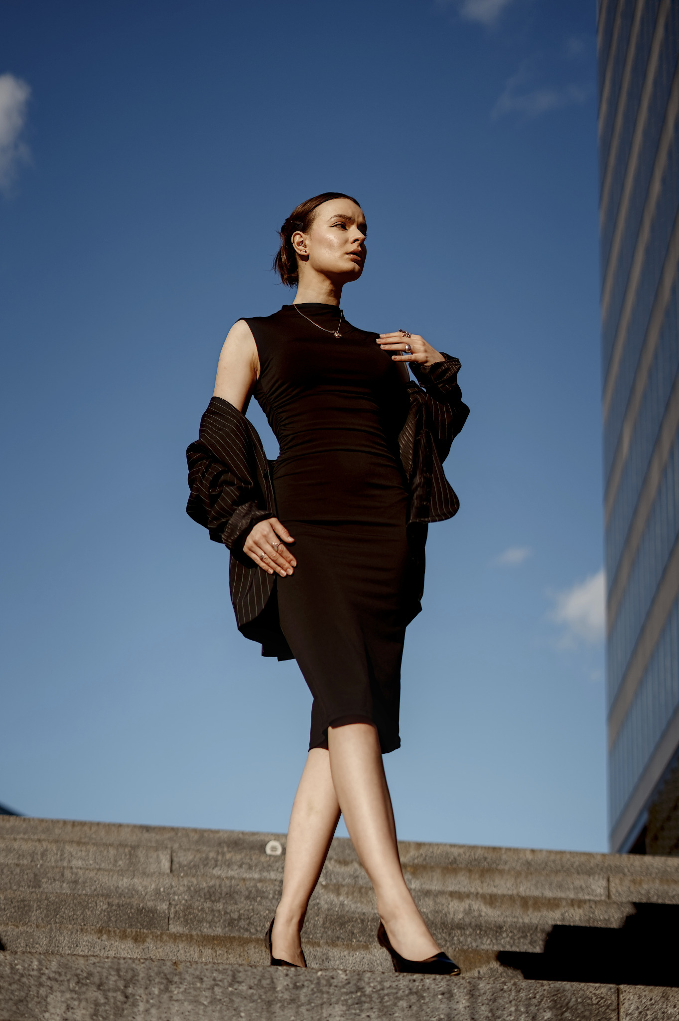 Elegancka kobieta w czarnej sukience i marynarce w paski pozuje na schodach, z nowoczesnym budynkiem w tle i błękitnym niebem. Ujęcie z dołu podkreśla sylwetkę.