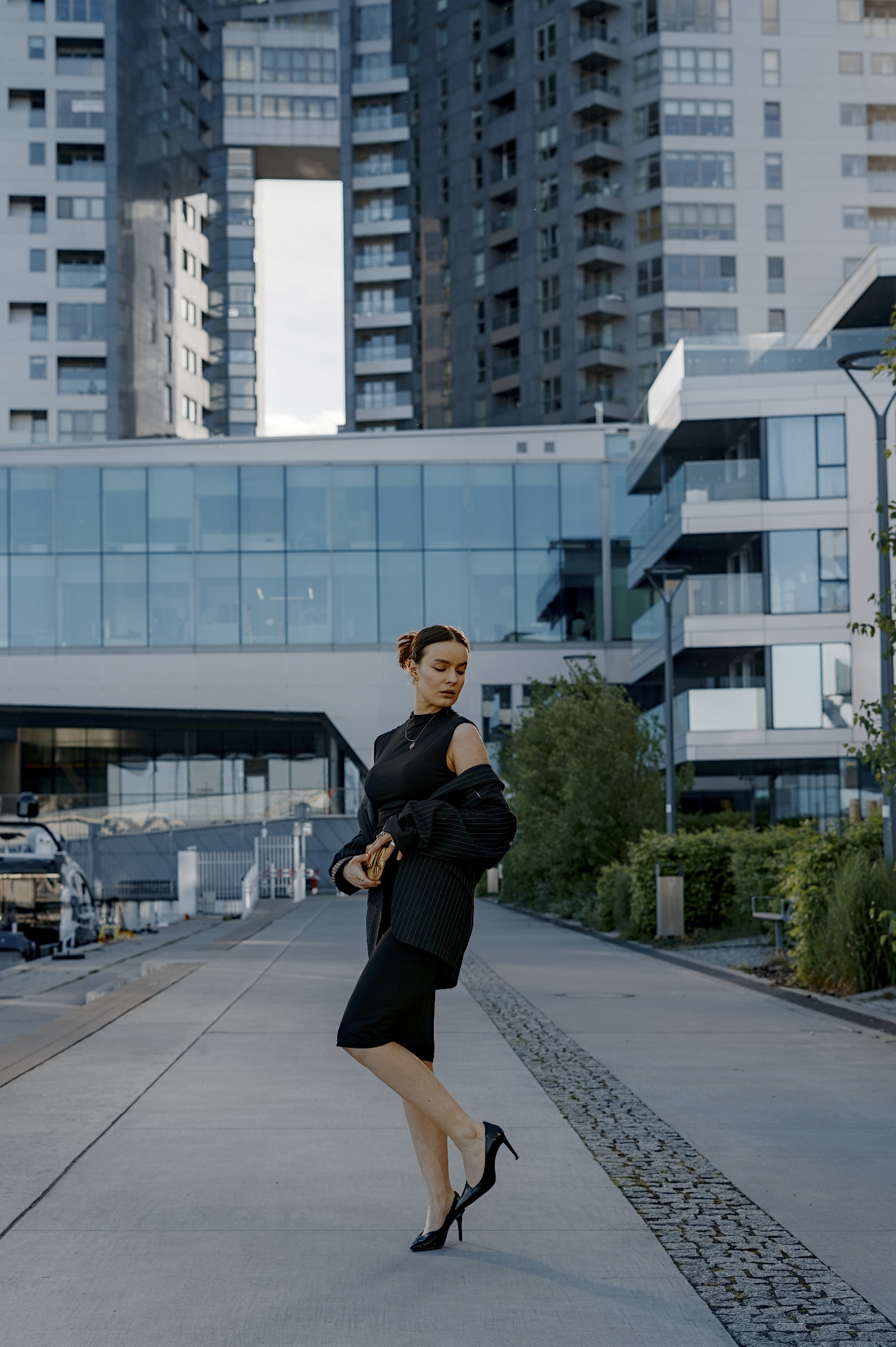 Elegancka kobieta w czerni pozuje na tle nowoczesnej architektury miejskiej. Uwagę przyciąga jej stylizacja i kompozycja zdjęcia z budynkami w tle.