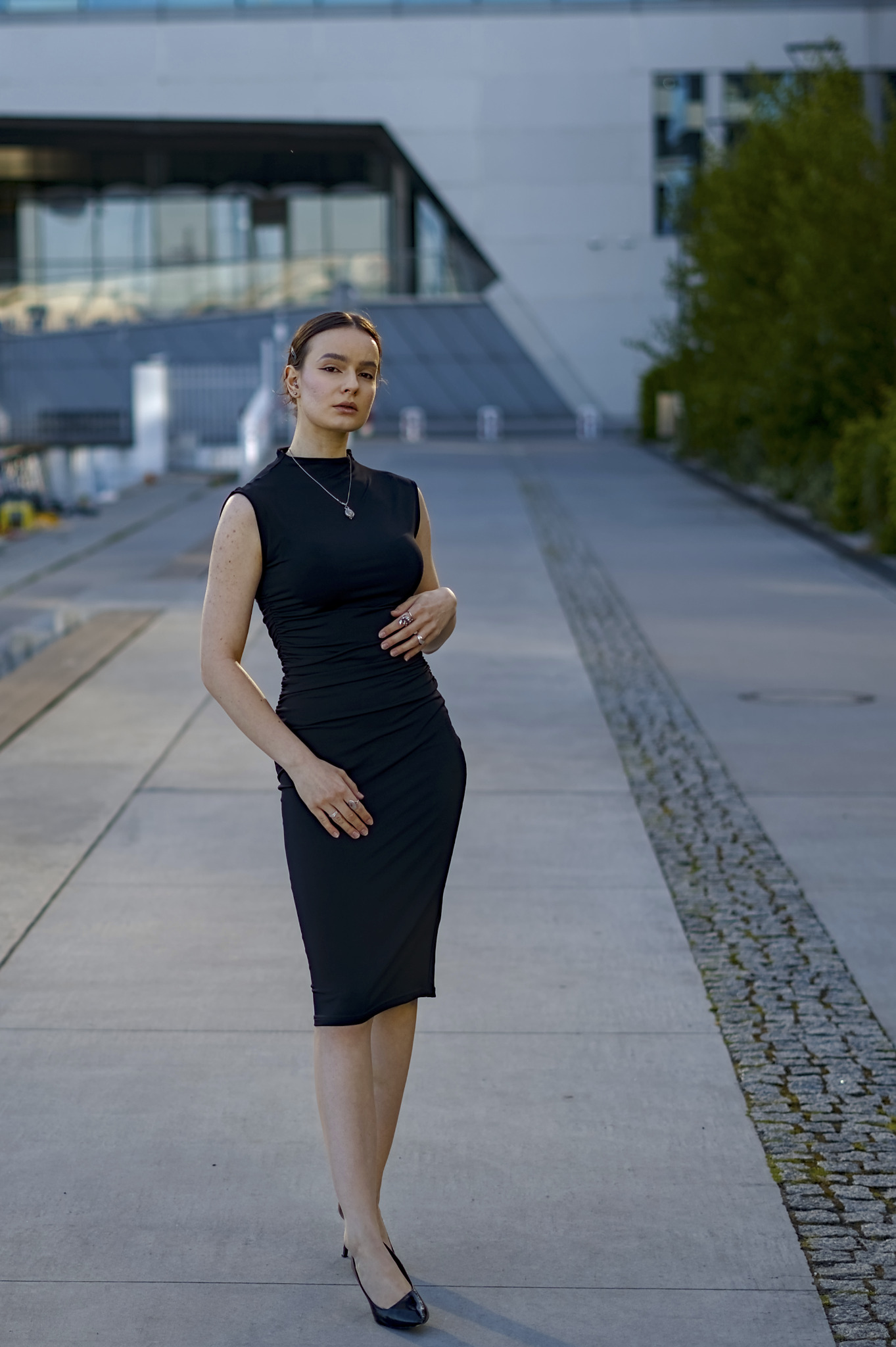 Elegancka kobieta w czarnej sukience pozuje na tle nowoczesnej architektury. Uwagę przykuwają subtelne detale biżuterii i wyrazisty makijaż.