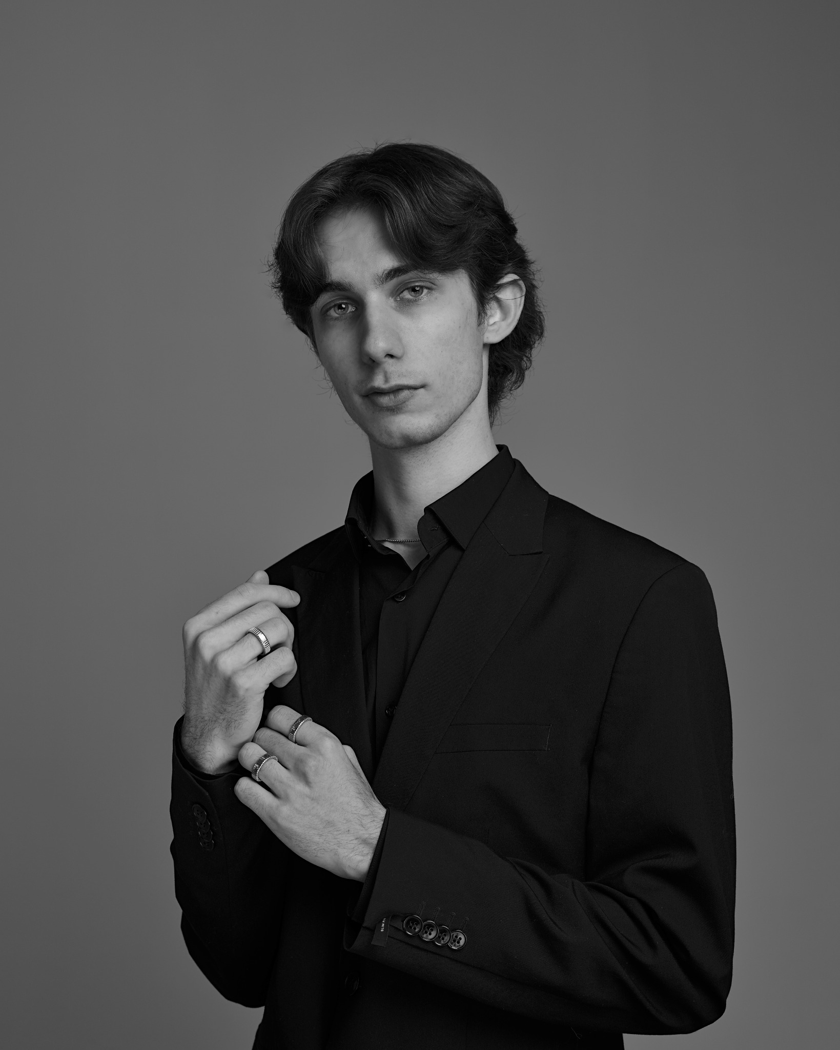 Czarno-biały portret młodego mężczyzny w czarnym garniturze, z pierścionkami na palcach, na jednolitym szarym tle. Ujęcie od pasa w górę, z delikatnym oświetleniem.