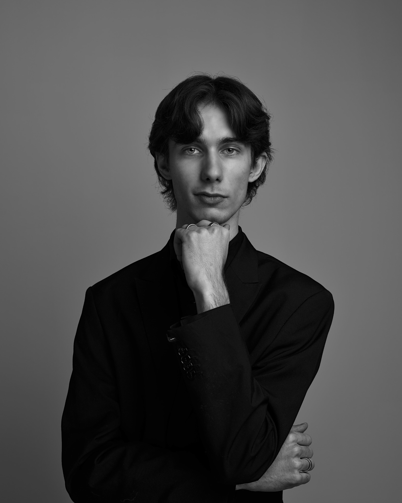 Czarno-biały portret młodego mężczyzny w czarnym garniturze, z ręką pod brodą, na jednolitym szarym tle. Ujęcie centralne, oświetlenie studyjne.