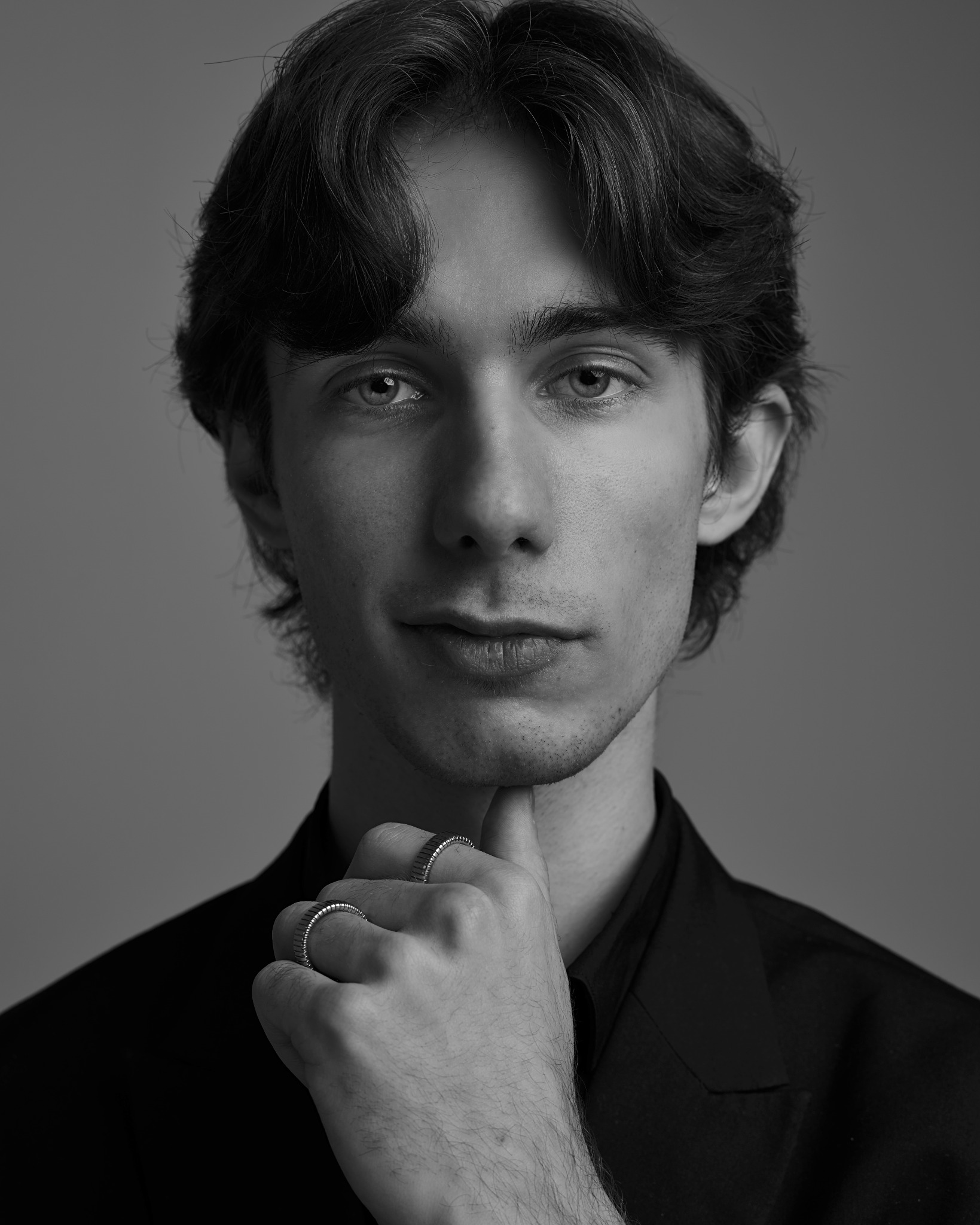 Czarno-biały portret młodego mężczyzny w garniturze, z ręką pod brodą i dwoma pierścieniami na palcach, na jednolitym tle. Ujęcie od przodu, skupione na twarzy i dłoniach.