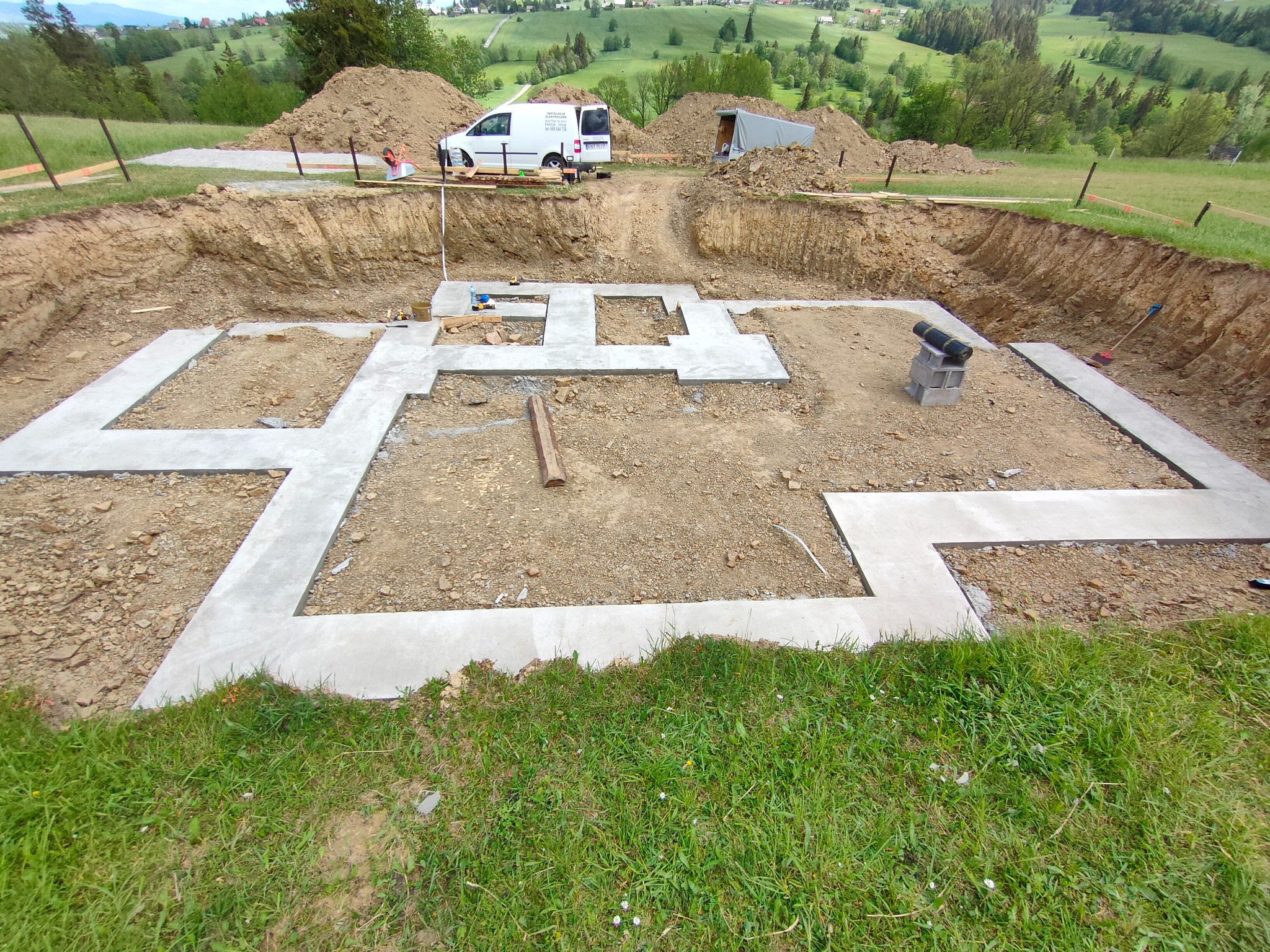 Wylane fundamenty domu na działce z wykopem, widok z góry. Na górze działki zaparkowany biały bus z reklamą firmy budowlanej. W tle zielone wzgórza.
