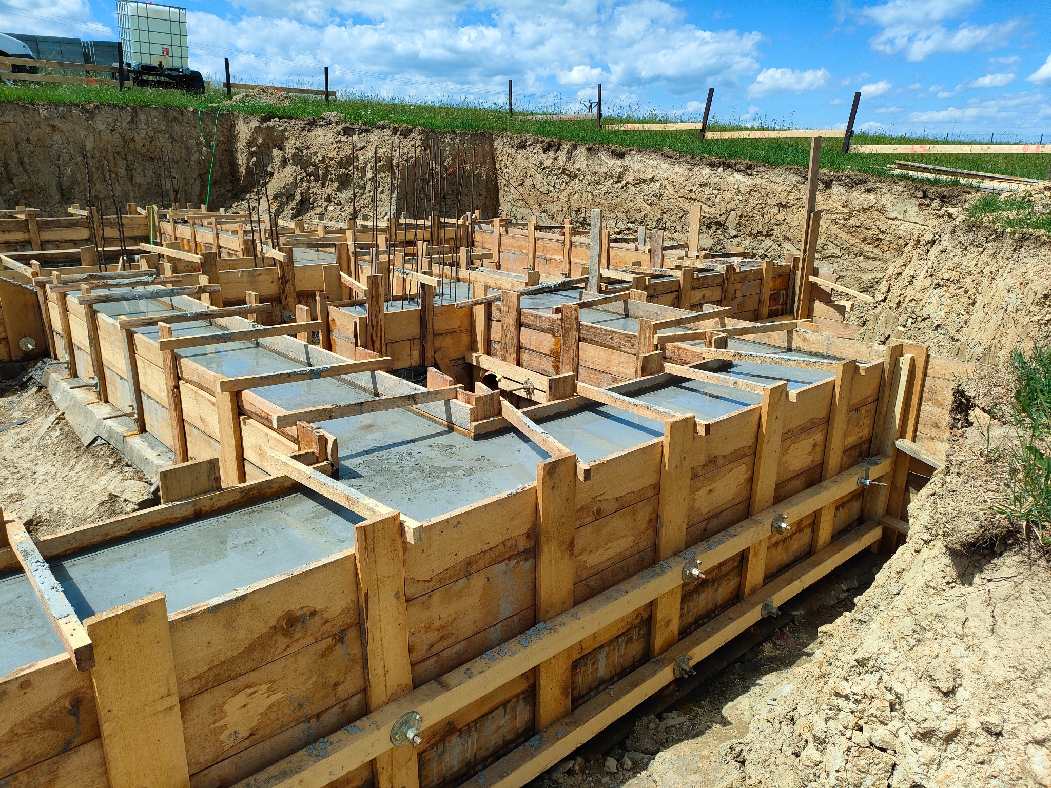 Zalane betonem drewniane szalunki fundamentów domu jednorodzinnego, widoczne zbrojenia i grunt. Słoneczny dzień, błękitne niebo z chmurami w tle.