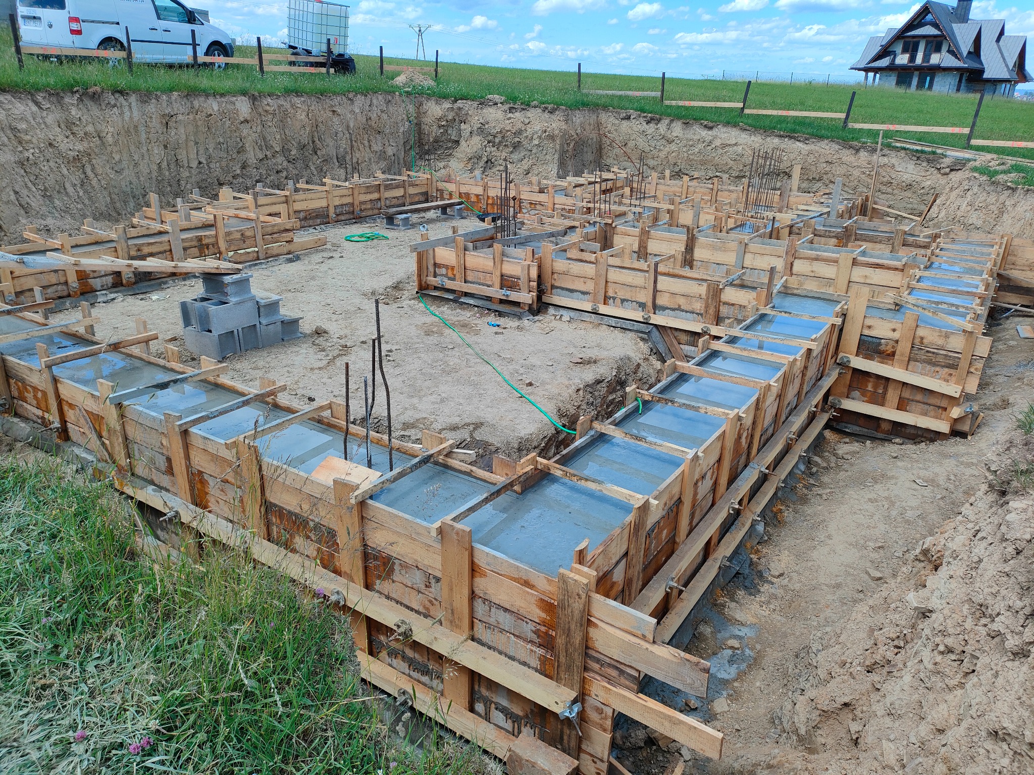 Zalane betonem szalunki fundamentowe na budowie domu jednorodzinnego, widok z góry. W tle ziemia, trawa i częściowo widoczny budynek w budowie.