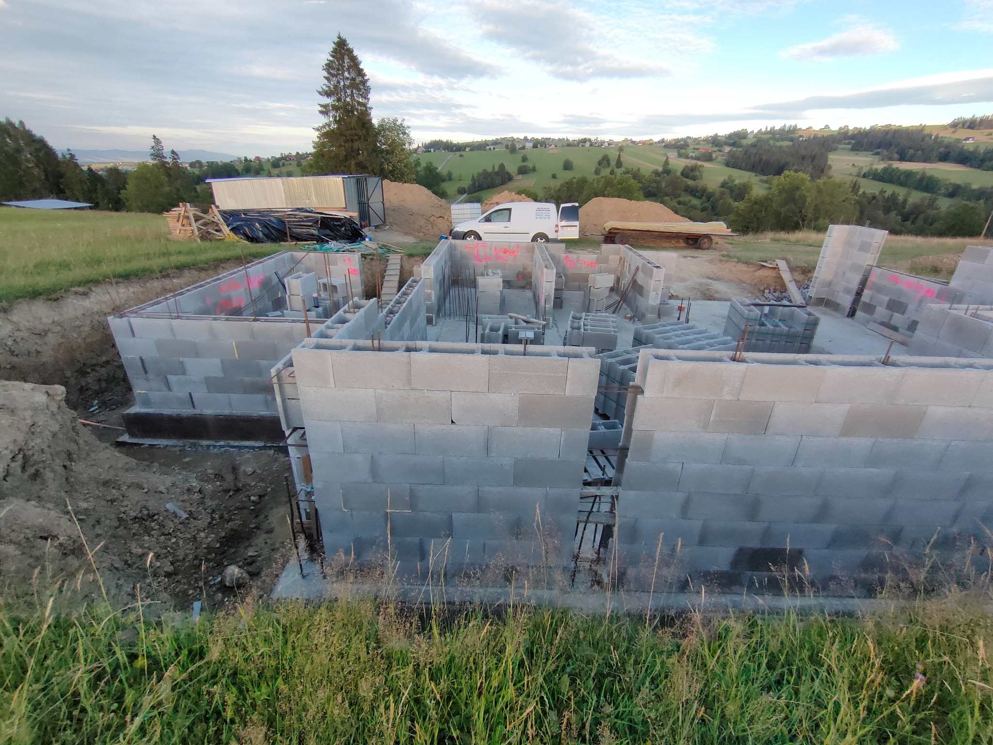 Budowa domu z bloczków betonowych na fundamencie, widok z góry na częściowo wzniesione ściany, w tle krajobraz wiejski i samochód dostawczy.