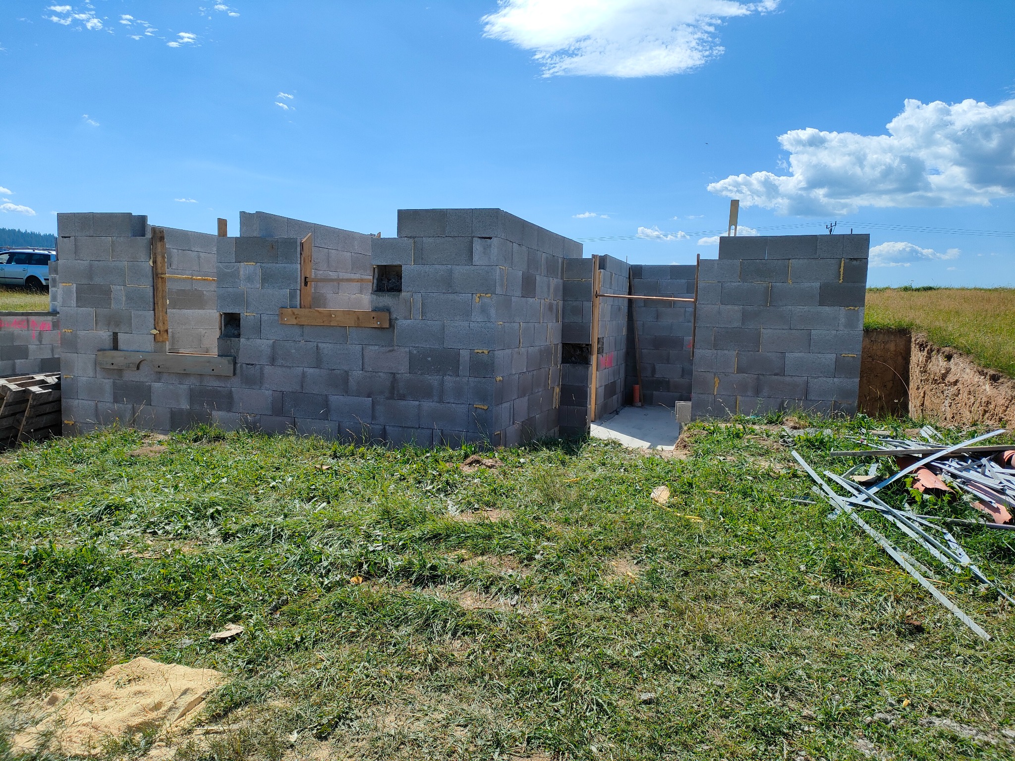 Wznoszenie ścian piwnicy z szarych bloczków betonowych na tle błękitnego nieba. Widoczne drewniane elementy szalunku i wykop ziemny.