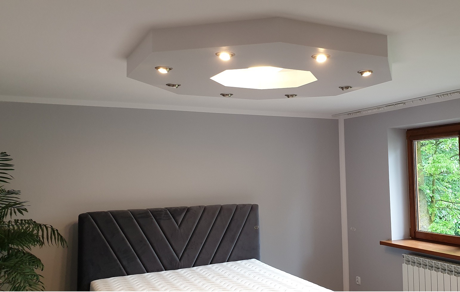 Sufit z zamontowanym, ośmiokątnym panelem oświetleniowym z halogenami i centralnym światłem w sypialni z szarymi ścianami i wezgłowiem łóżka.