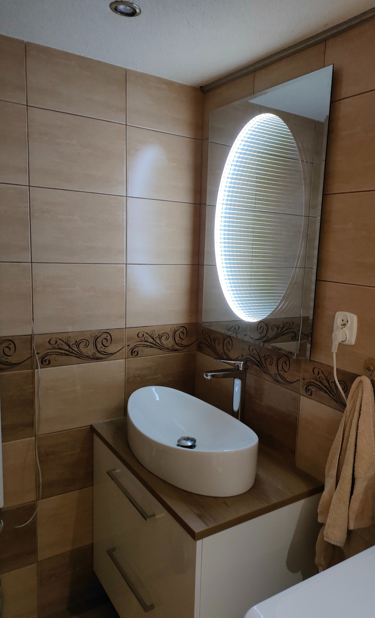 Nowoczesna łazienka z owalną umywalką nablatową, chromowaną baterią i podświetlanym lustrem 3D na tle beżowych płytek z dekoracyjnym motywem.