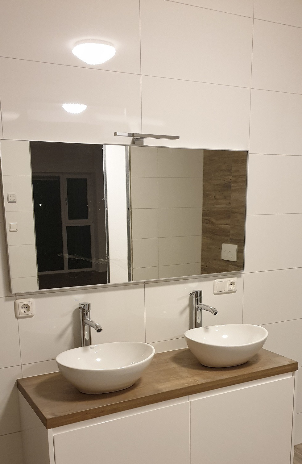 Nowoczesna łazienka z dwoma umywalkami nablatowymi, lustrem z oświetleniem LED i białymi płytkami. Widoczne gniazdka elektryczne i armatura.