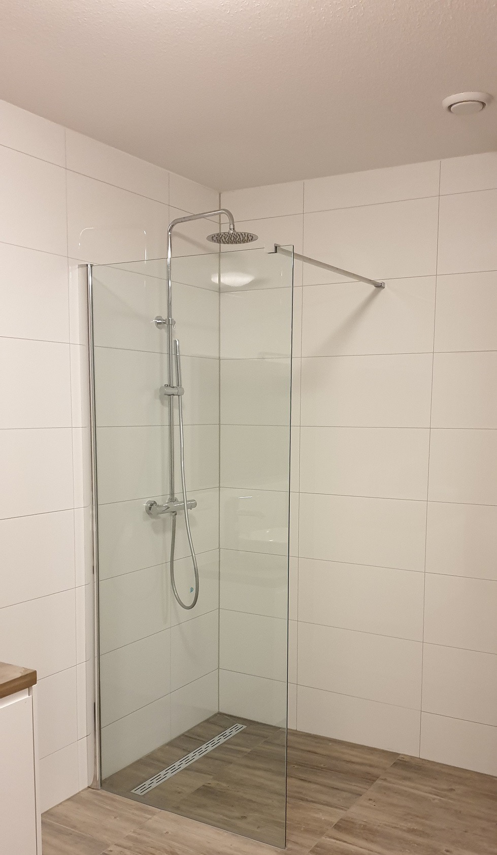 Nowoczesna kabina prysznicowa z chromowaną armaturą i odpływem liniowym, wykończona białymi płytkami i drewnopodobną podłogą. Minimalistyczny design.