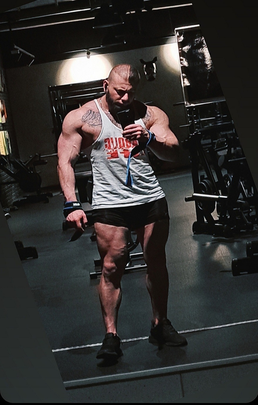 Mężczyzna z tatuażami, w sportowej koszulce i spodenkach, robi sobie selfie w siłowni, prezentując imponującą muskulaturę i żyły na nogach.
