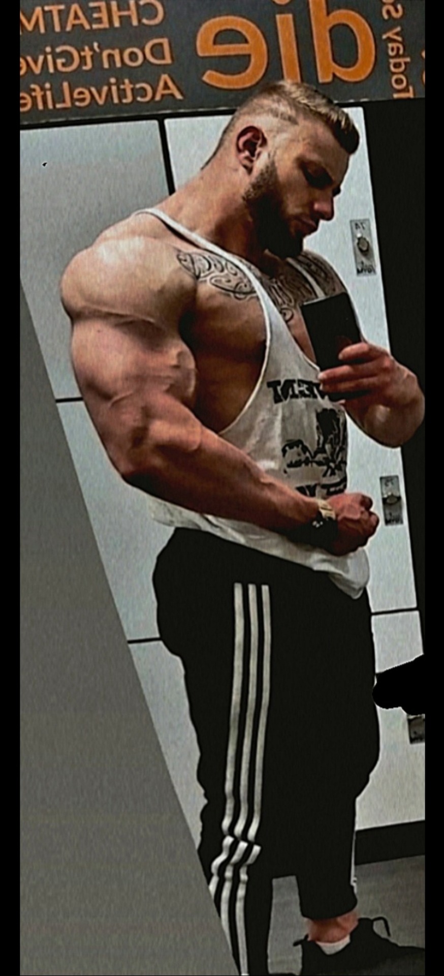 Mężczyzna z tatuażami prezentuje muskulaturę w lustrze szatni w Chorzowie, ubrany w tank top i spodnie dresowe z trzema paskami.
