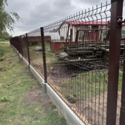 Nowo zainstalowane, brązowe ogrodzenie z paneli z betonową podmur&oacute;wką na trawiastym terenie, widoczne słupki i elementy mocujące, w tle budynek gospodarczy i przyczepa.