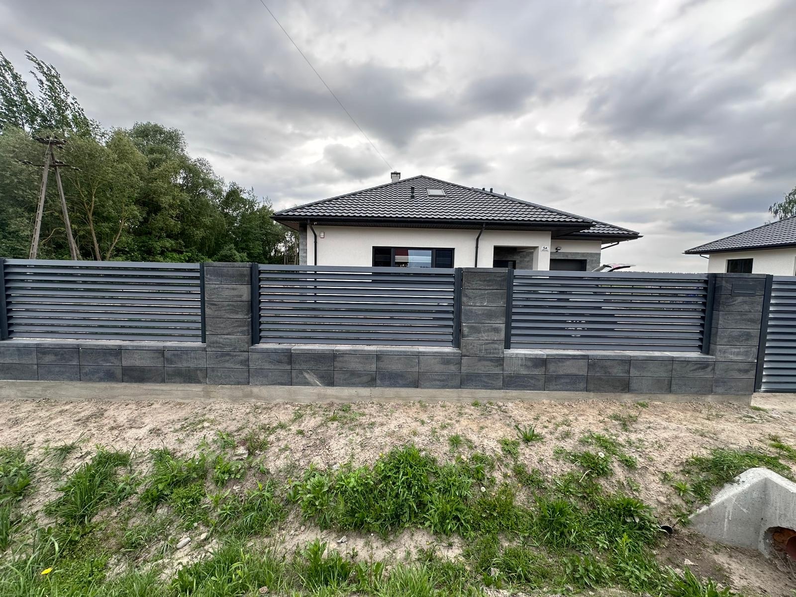 Nowoczesne ogrodzenie z poziomych, szarych paneli metalowych i betonowych słupków, na tle domu z ciemną dachówką i pochmurnego nieba.