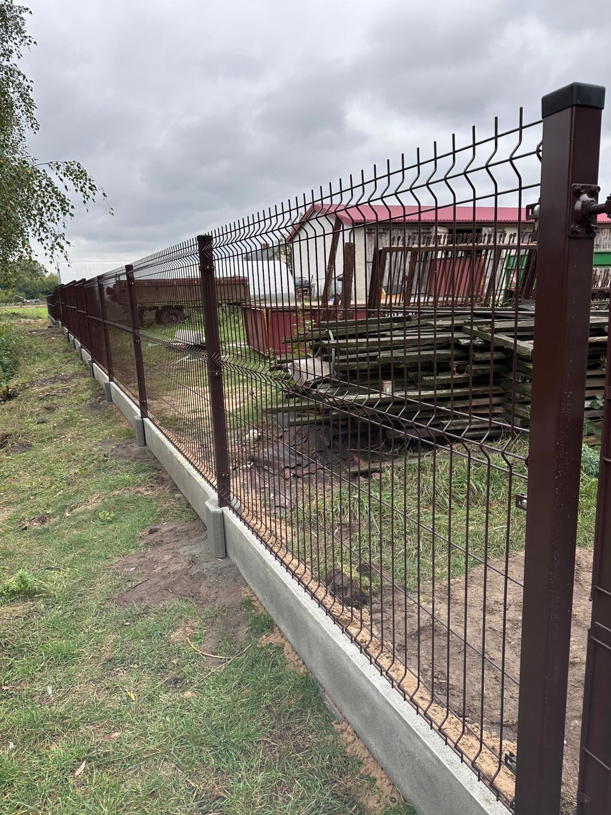 Nowo zainstalowane, brązowe ogrodzenie z paneli z betonową podmurówką na trawiastym terenie, widoczne słupki i elementy mocujące, w tle budynek gospodarczy i przyczepa.