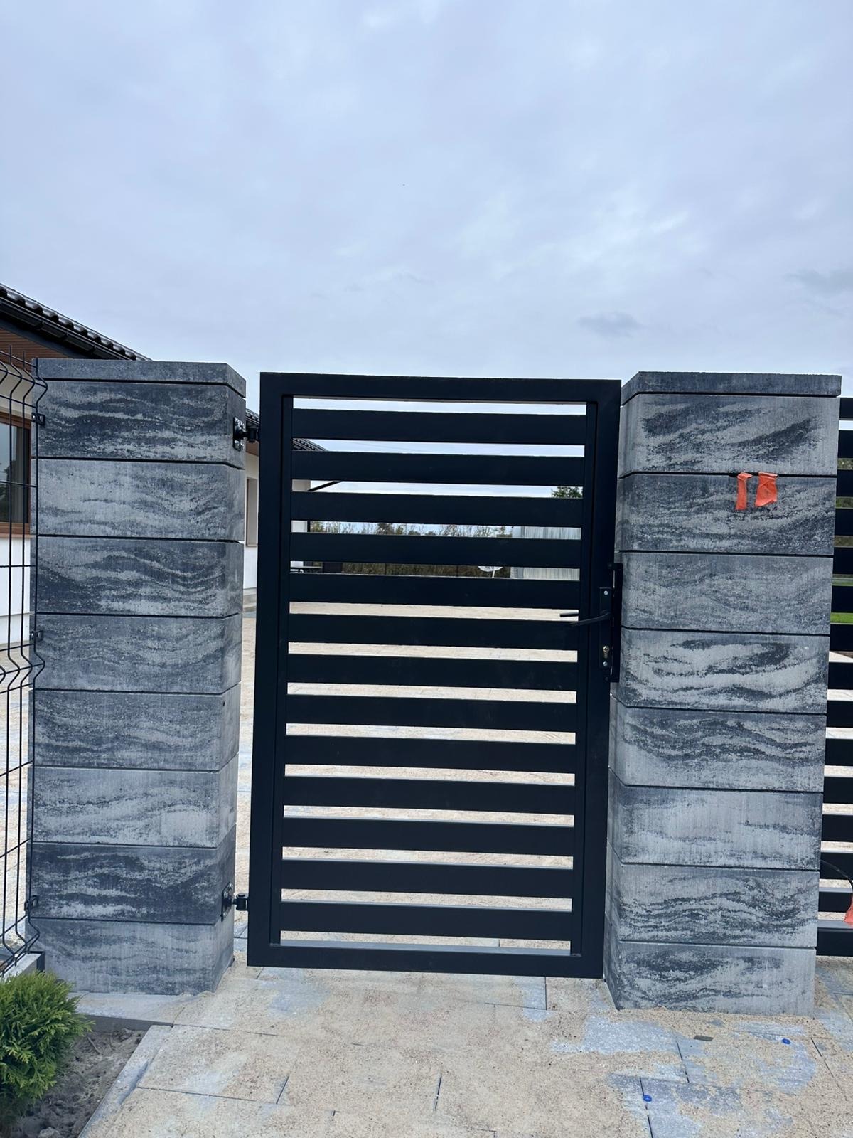 Nowoczesna brama wjazdowa z poziomymi, czarnymi profilami, osadzona na słupkach z szarej kostki imitującej kamień, widok z zewnątrz na tle pochmurnego nieba.