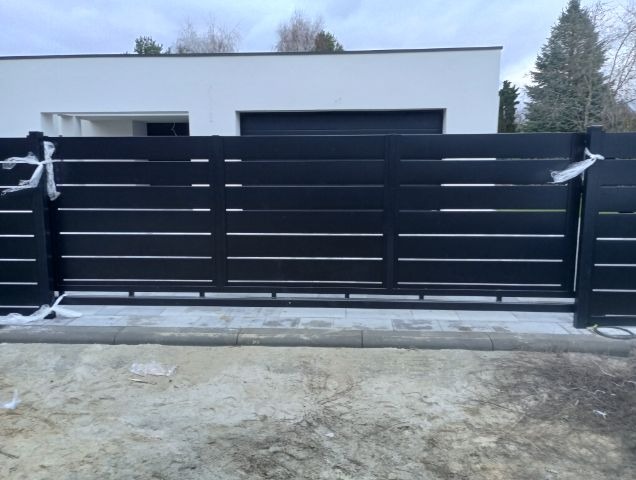 Nowoczesna, czarna brama przesuwna z poziomymi szczeblami, częściowo jeszcze zabezpieczona folią ochronną, na tle białego domu z garażem.