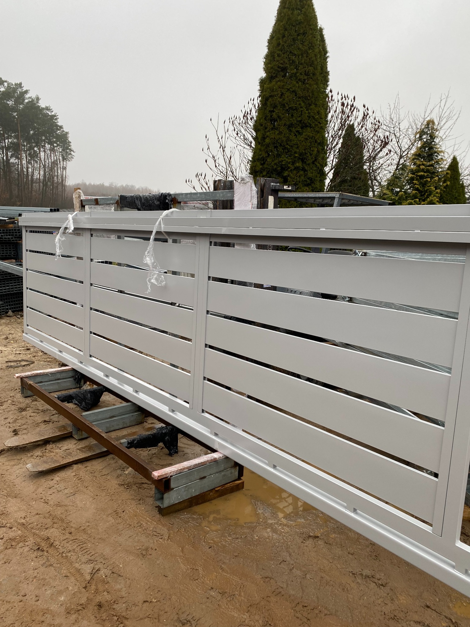 Biała, przesuwna brama metalowa z poziomymi panelami, częściowo owinięta folią ochronną, umieszczona na metalowej konstrukcji na błotnistym terenie, w tle drzewa i pochmurne niebo.