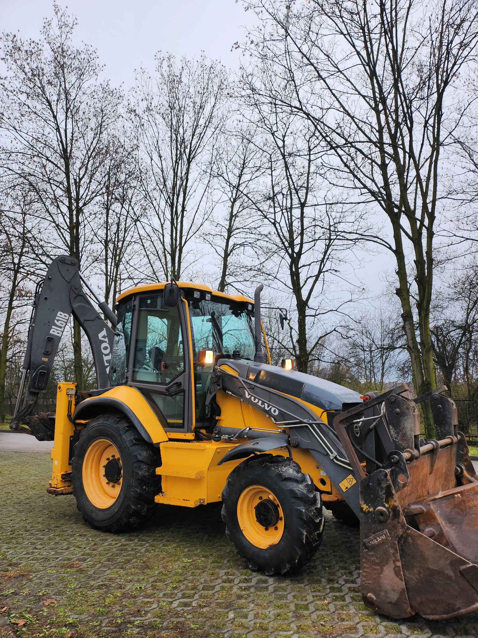 Żółta koparko-ładowarka Volvo stoi na kostce brukowej, z widocznymi drzewami bez liści w tle, pochmurne niebo.