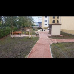 Budownictwo Drogowe Brukarstwo Robert Wieczorek - Czerwona kostka brukowa tworzy ścieżkę wzdłuż trawnika z młodymi drzewkami i ławkami. W tle budynek i pracownik z taczką. Widoczny fragment węża ogrodowego.