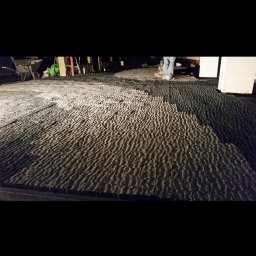 Budownictwo Drogowe Brukarstwo Robert Wieczorek - Ułożona, szara kostka brukowa o nieregularnej fakturze, widok z dołu, z fragmentem osoby w tle. Nierówne oświetlenie podkreśla strukturę materiału.