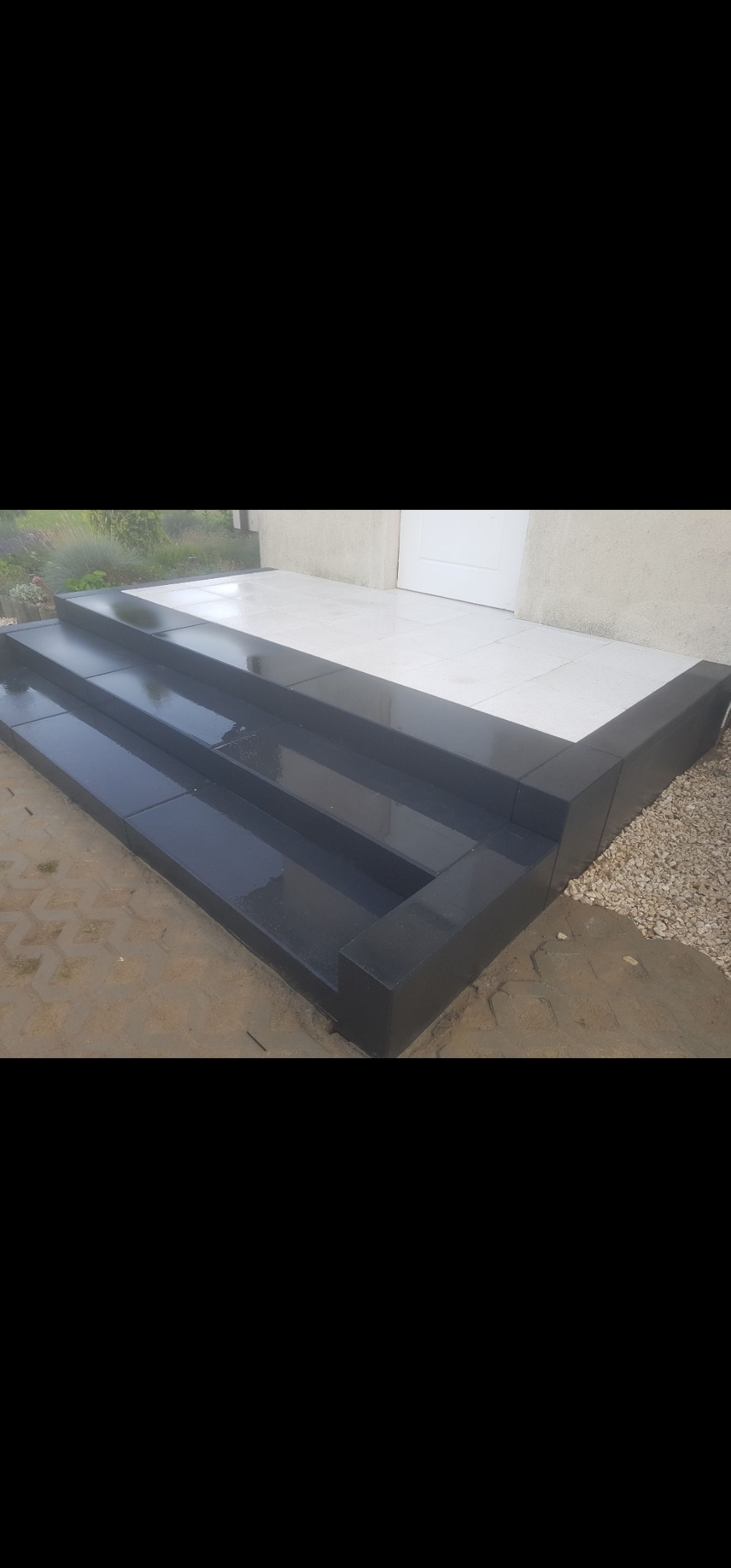 Nowoczesne, czarne schody z kostki brukowej prowadzące do jasnego wejścia domu. Widoczne detale wykończenia i otoczenie z kamyków.