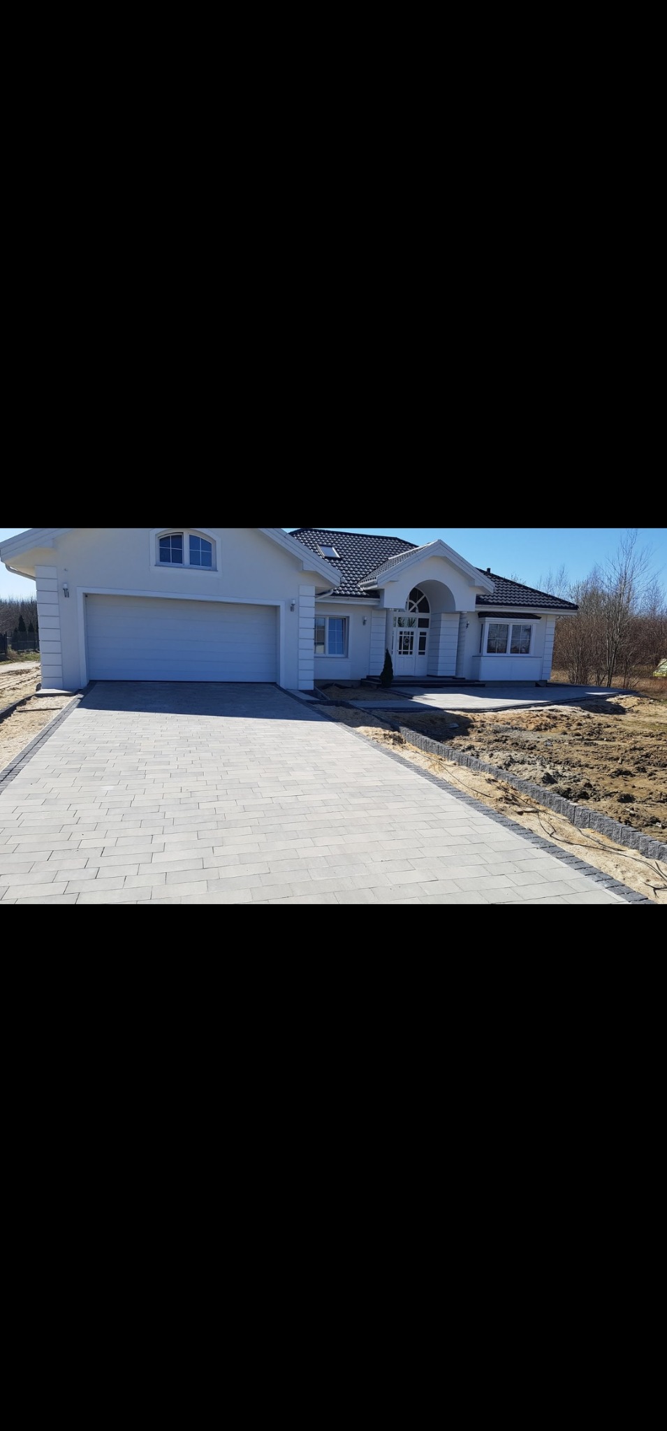 Jasnoszara kostka brukowa prowadząca do białego domu z garażem. Widoczne obrzeża z ciemnej kostki. Częściowo przygotowany teren pod ogród.