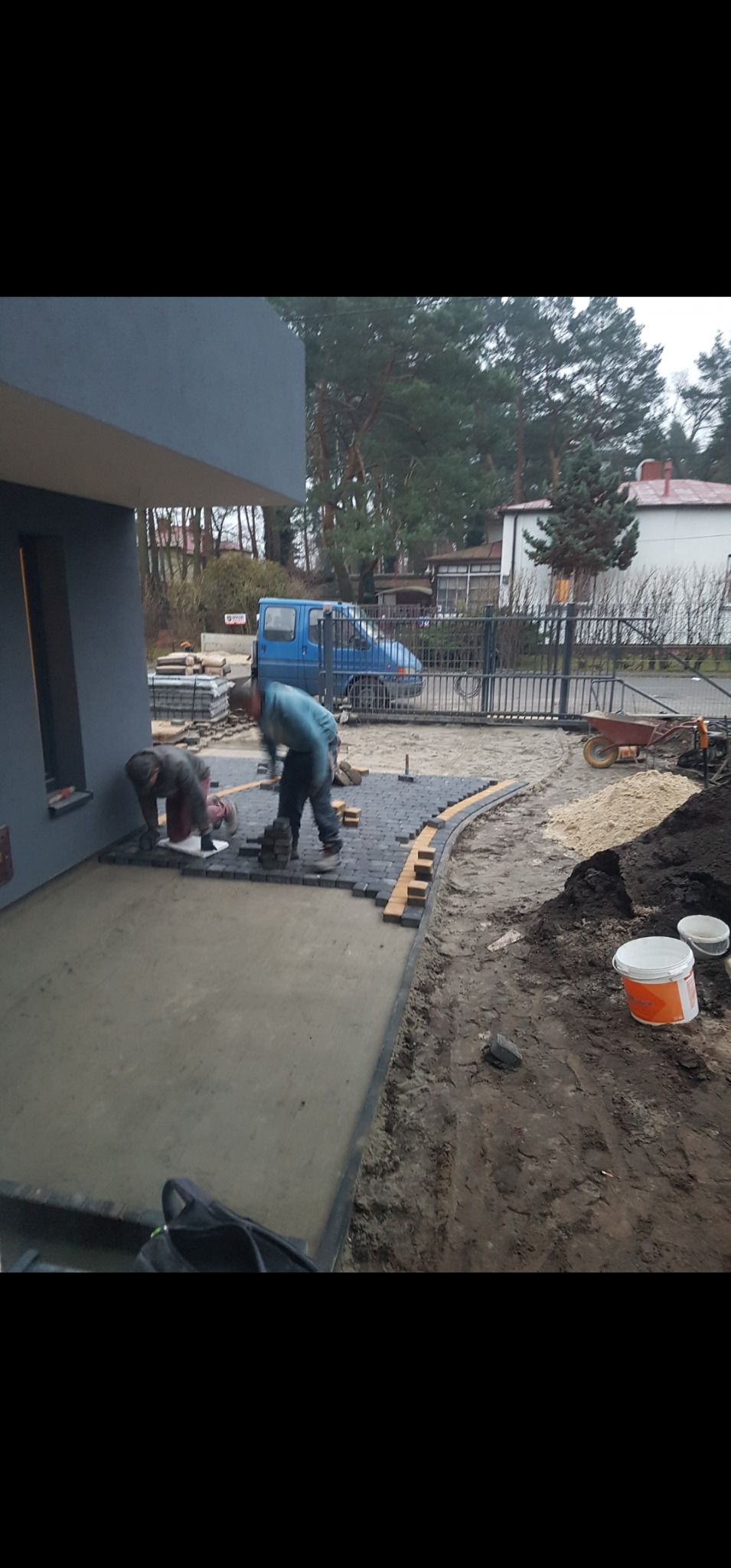 Dwóch pracowników układa kostkę brukową obok domu z szarą elewacją. Częściowo ułożona kostka, widoczne narzędzia i materiały budowlane. W tle zaparkowany niebieski bus.