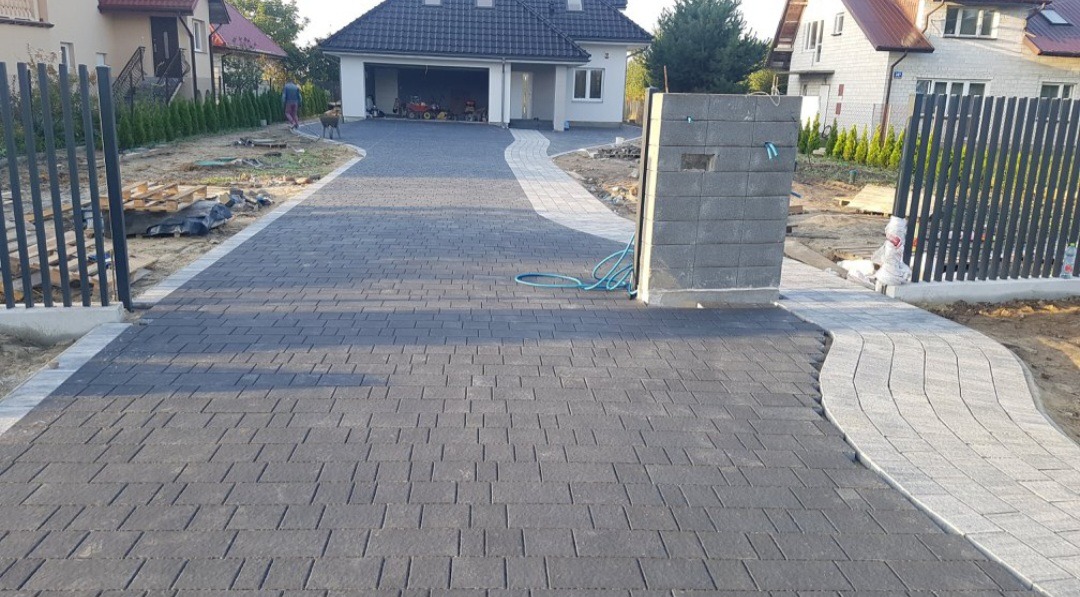 Ułożona, grafitowa kostka brukowa na podjeździe do domu z garażem. Jasnoszara kostka wyznacza ścieżkę. Częściowo postawiony murek z szarego bloczka.