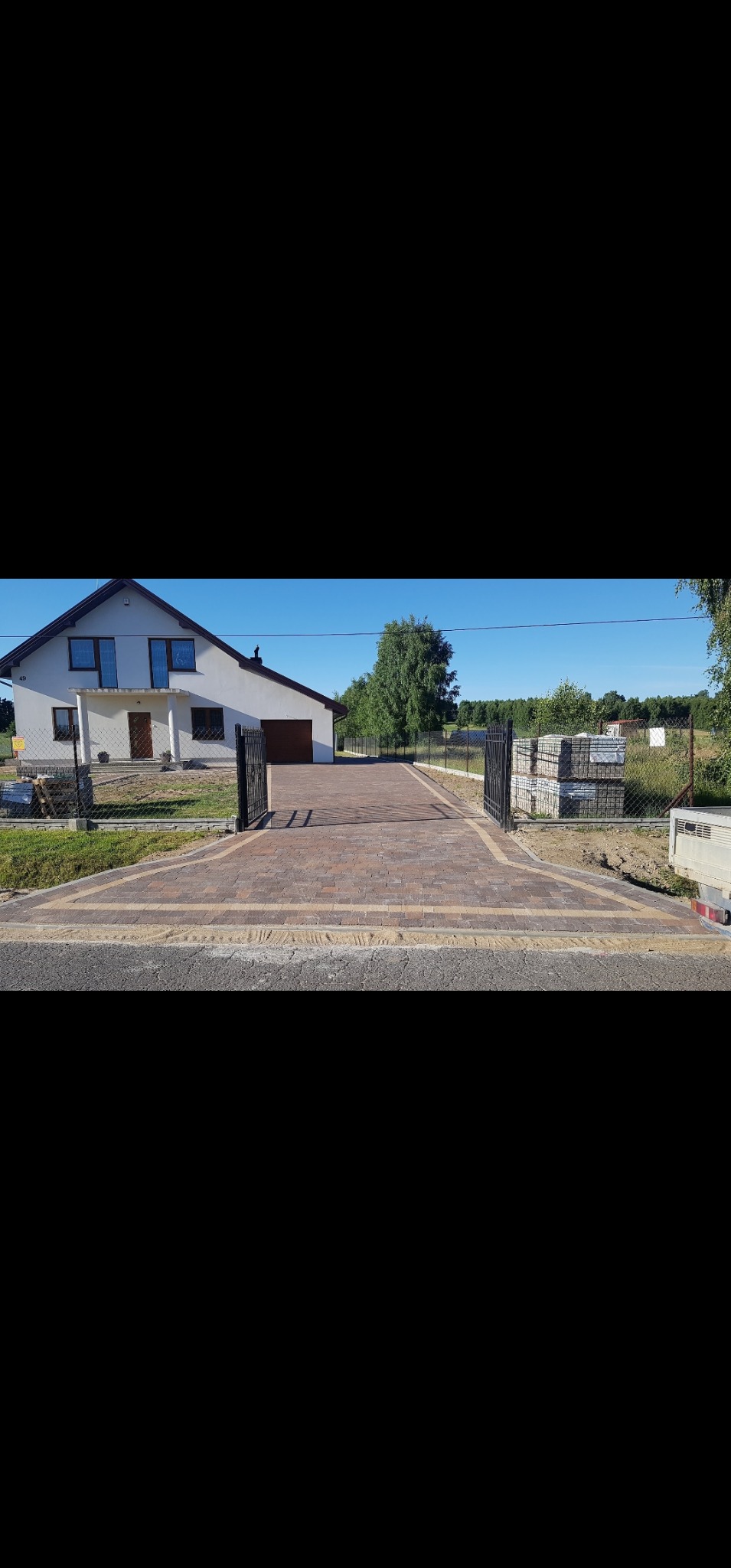 Ułożona kostka brukowa w odcieniach brązu i beżu tworzy wzór na podjeździe przed domem z otwartą, czarną bramą. W tle widoczne palety z kostką i zieleń.