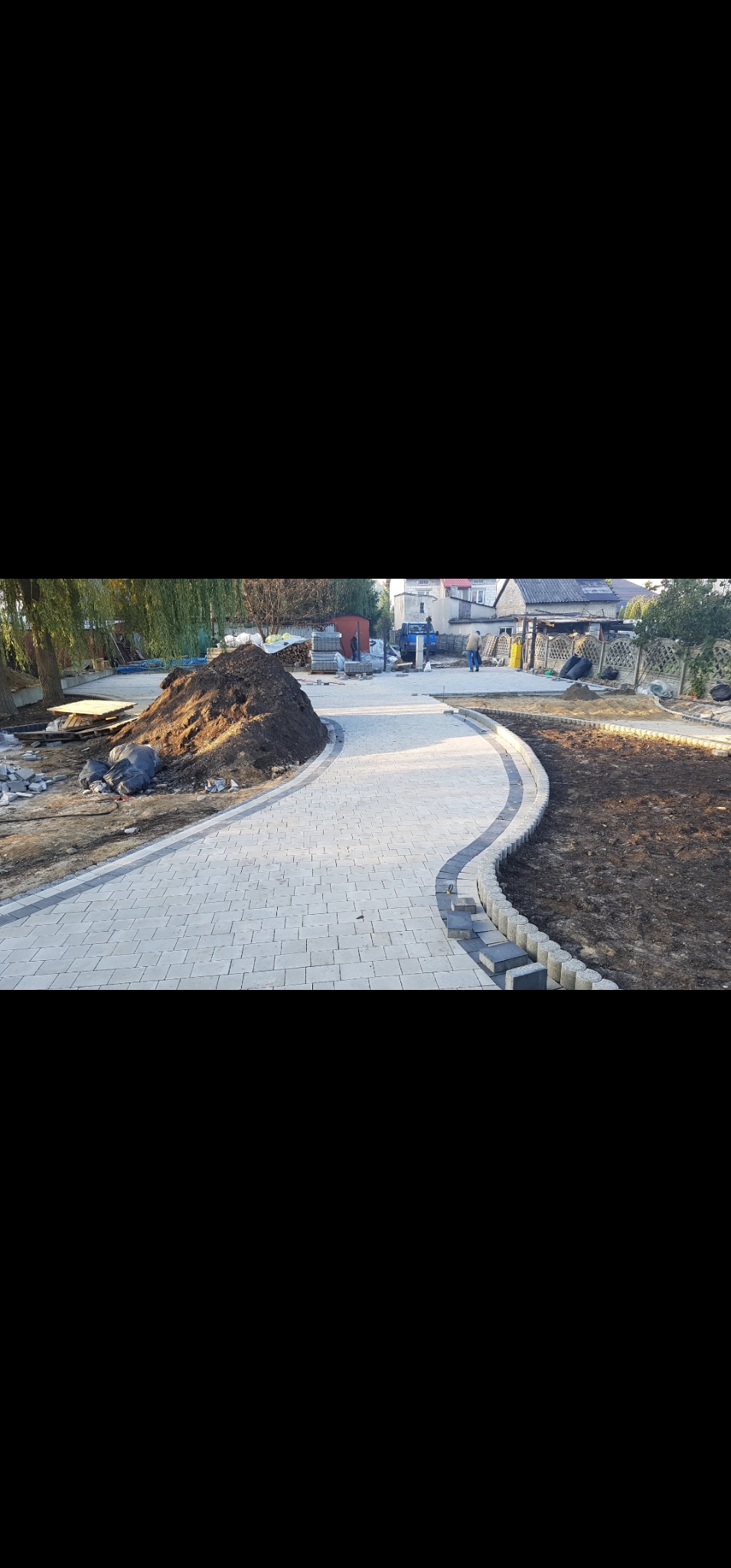 Ścieżka z jasnoszarej kostki brukowej w ogrodzie, z widoczną stertą ziemi i elementami przygotowania terenu. Częściowo ukończona realizacja.