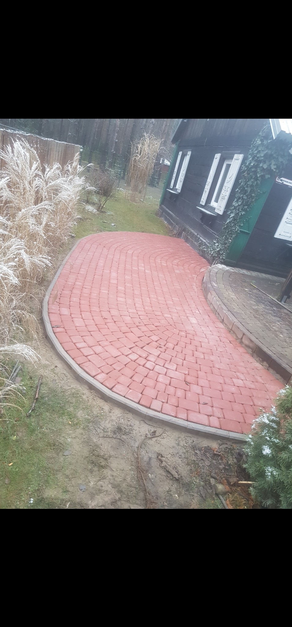 Czerwona kostka brukowa ułożona w łukowaty chodnik prowadzący do domu z zielonymi pnączami. Szara obwódka, trawa i rośliny w tle. Zimowa sceneria.
