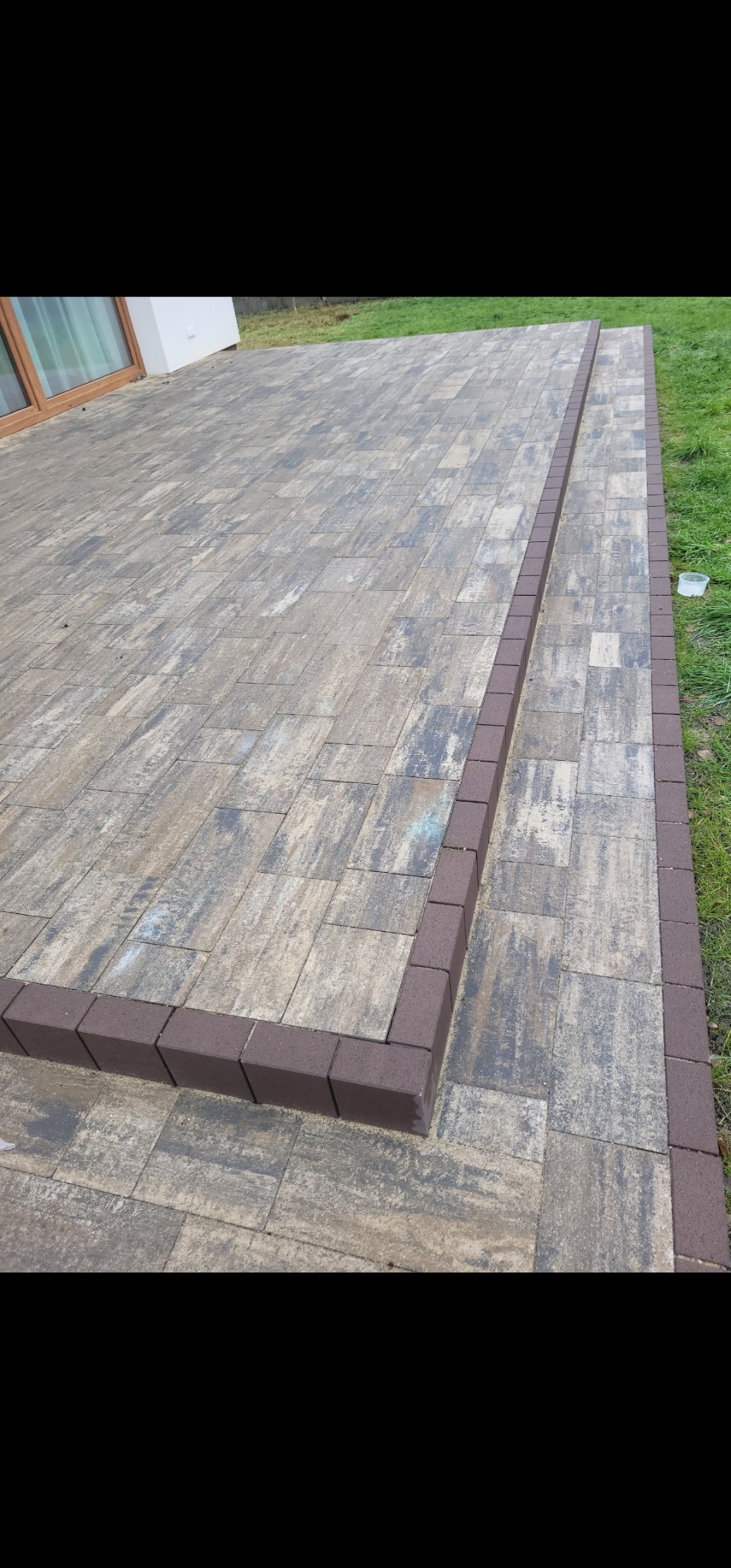 Ułożona kostka brukowa w odcieniach szarości i beżu, obramowana ciemnobrązową kostką. Widoczny fragment budynku i trawnika. Układanie kostki z wyraźnym wzorem.