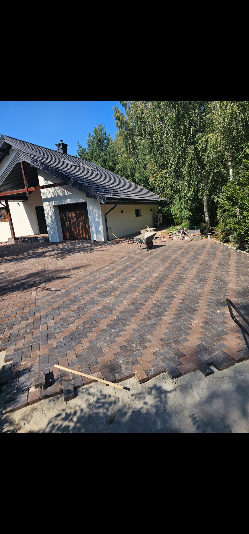 Dwukolorowa kostka brukowa ułożona w jodełkę na podjeździe domu jednorodzinnego, widoczne narzędzia i piasek, etap prac wykończeniowych, słoneczny dzień.