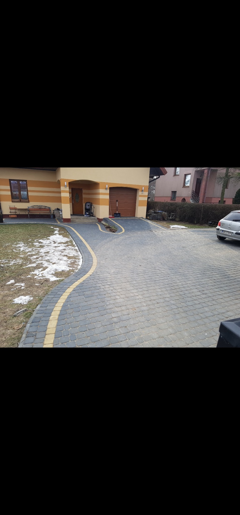 Ułożona szara kostka brukowa z żółtym obramowaniem na podjeździe do domu jednorodzinnego. Częściowo stopniały śnieg na trawniku. Widoczny samochód osobowy.