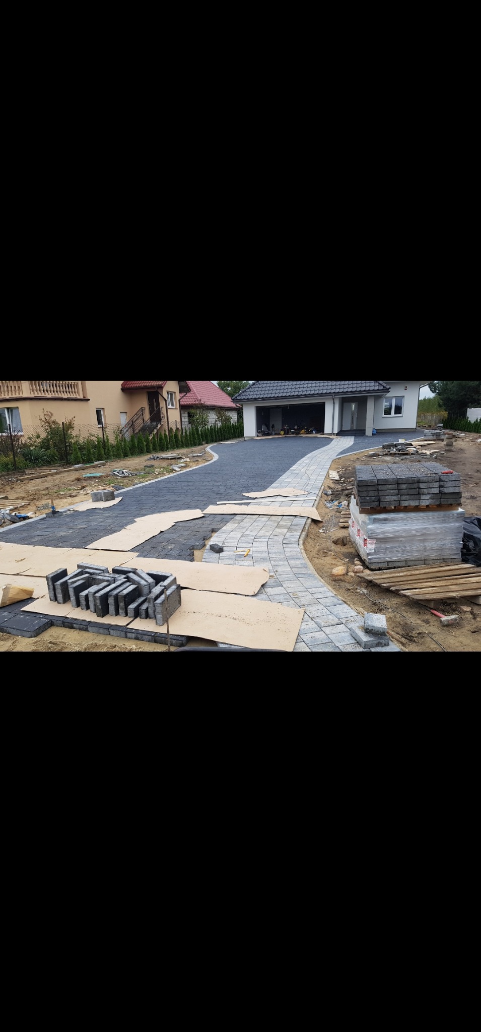 Fragment układania kostki brukowej przed domem jednorodzinnym. Część nawierzchni ułożona z szarej i grafitowej kostki, widoczne palety z kostką i kartony zabezpieczające.