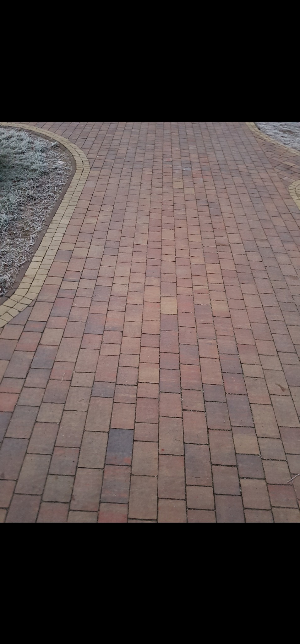 Ułożona kostka brukowa w odcieniach czerwieni i brązu, obrzeże z jasnej kostki. Na obrzeżach trawa pokryta szronem. Widok z perspektywy.
