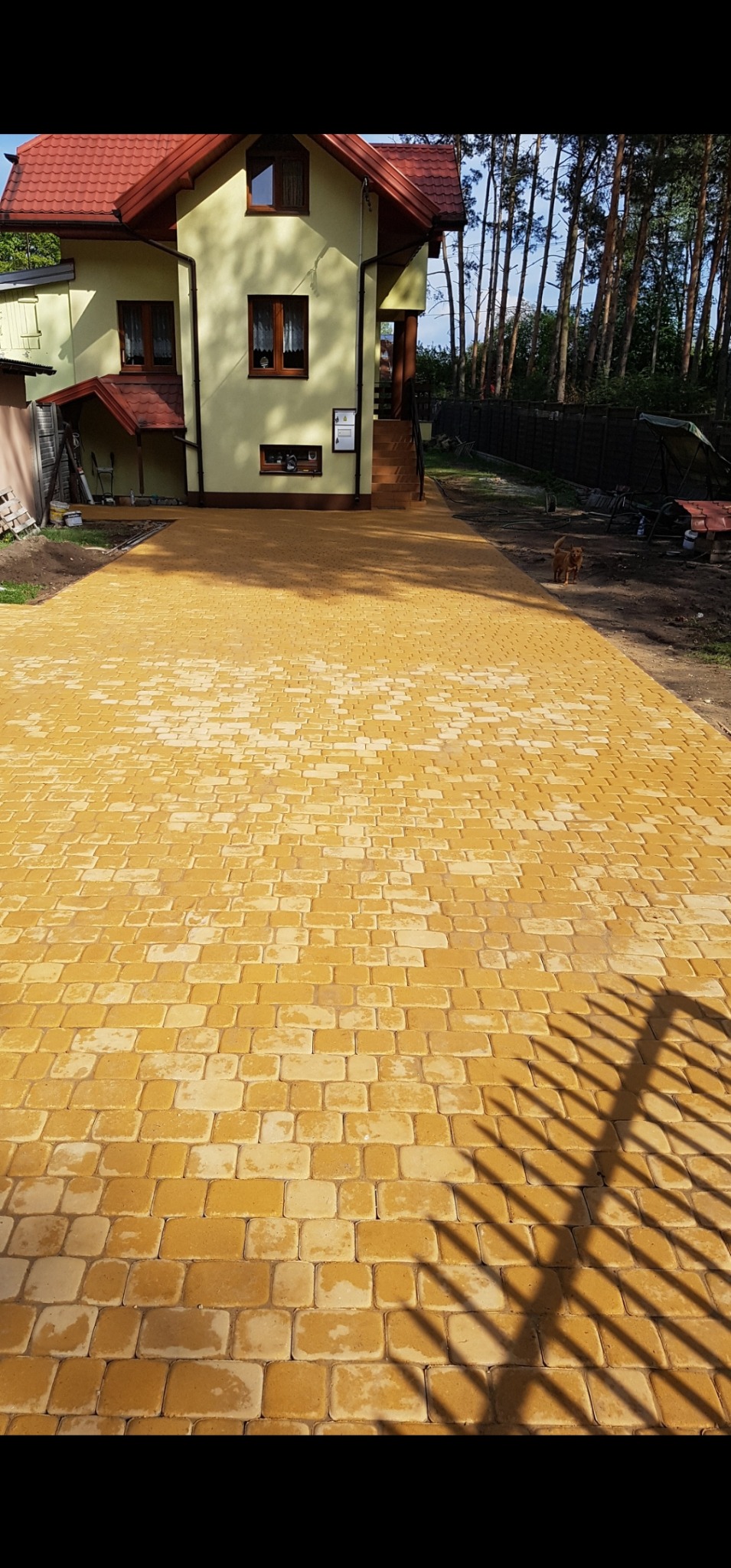 Żółta kostka brukowa prowadząca do domu z czerwonym dachem, w tle drzewa. Cień ogrodzenia na kostce. Widok na posesję po wykonanej usłudze.
