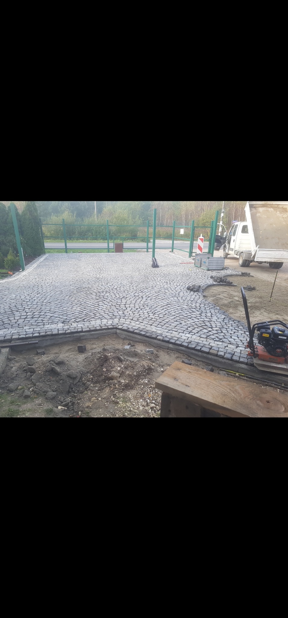Układanie kostki granitowej na podjeździe, widoczny fragment placu z ułożoną kostką, obrzeża i przygotowany teren pod dalsze prace. W tle brama i samochód ciężarowy.