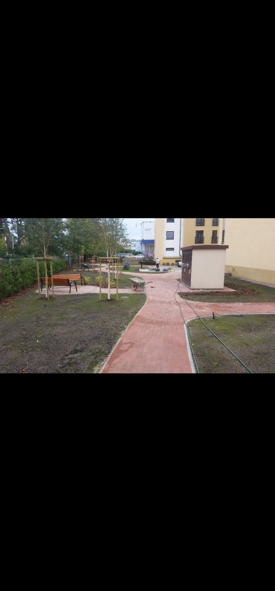 Czerwona kostka brukowa tworzy ścieżkę wzdłuż trawnika z młodymi drzewkami i ławkami. W tle budynek i pracownik z taczką. Widoczny fragment węża ogrodowego.