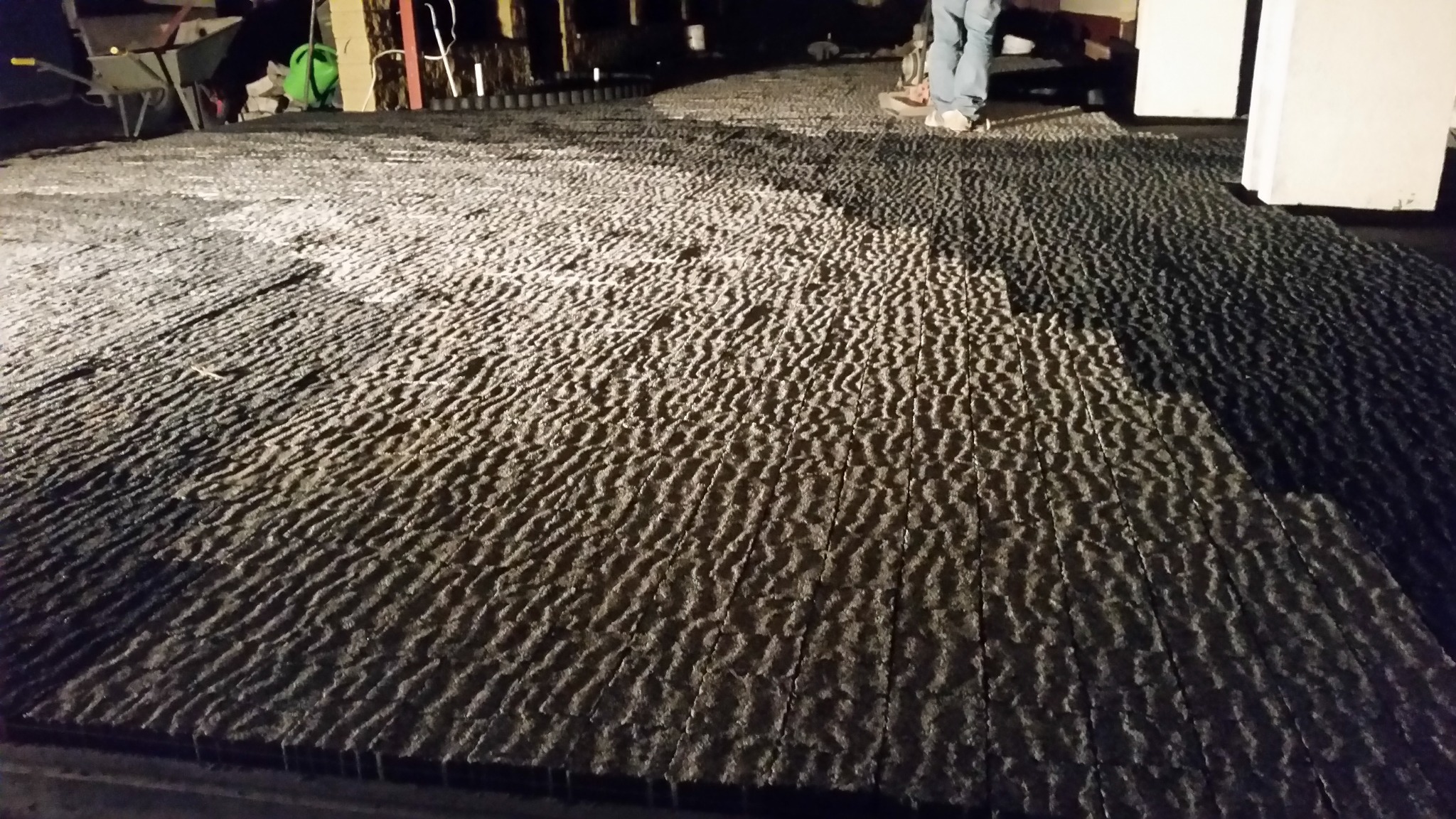 Ułożona, szara kostka brukowa o nieregularnej fakturze, widoczna z niskiego kąta. W tle fragment osoby i narzędzia budowlane. Oświetlenie sztuczne.